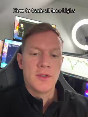 A man with fair skin is centered in the frame, looking towards the viewer and speaking. He has short, light brown hair and is wearing a black shirt. Behind him, multiple computer monitors display charts and graphs, illuminated by screen light and a ring light on the right side.