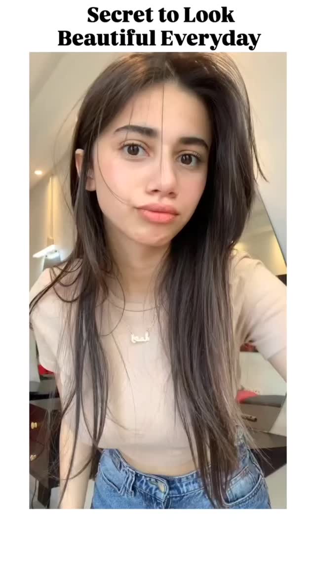 The image shows a woman with long, wavy dark hair, divided into two halves by a vertical line drawn down the middle. Her face is centered, and she's looking towards the camera with a slight pout. She's wearing a light-colored top and jeans. The background is blurred, and it seems to be a mirror and bright lighting.