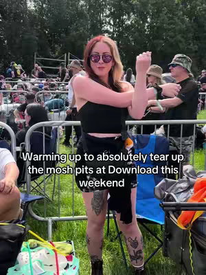 A woman in sunglasses and a black top is standing outdoors at a music festival, stretching her arms. The background shows a crowd of people, metal barriers, and green grass. Text overlayed on the image reads 'Warming up to absolutely tear up the mosh pits at Download this weekend'.