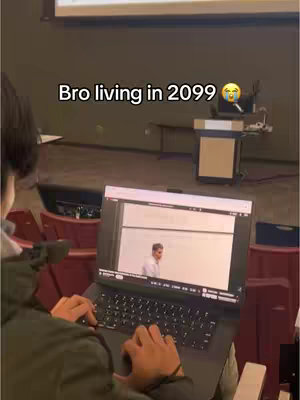 A person is sitting in a lecture hall using a laptop. The laptop screen displays a presentation with text and a man in a white shirt. The hook text 'Bro living in 2099 😭' is overlaid, creating a humorous effect.