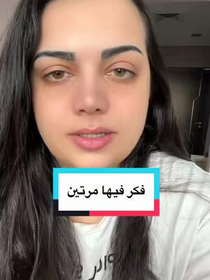 A woman appears in the foreground, centered, against an out-of-focus background. She faces the viewer directly with a neutral expression. The foreground features a rectangular text overlay in the center of the screen, containing Arabic text on a colored background. She has long, dark hair and is wearing a light-colored top, indicating a casual setting. The lighting seems natural.