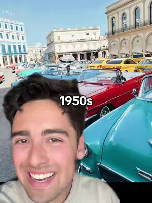 A selfie-style shot with a man smiling in the foreground set against a background of vintage cars parked on a street in Cuba. The man is smiling at the camera and there is text overlayed on the image, displaying the text "1950s". The background is filled with blue, red, and yellow convertibles, with various buildings.