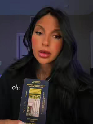 A woman with long dark hair and makeup holds up a tarot card, looking towards the camera in a room with dim lighting.