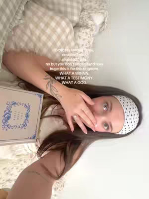 A woman wearing a white headband holding a Bible in bed, propping herself up with her arm. The woman is looking at the camera, with one hand near her mouth. The image is rotated.