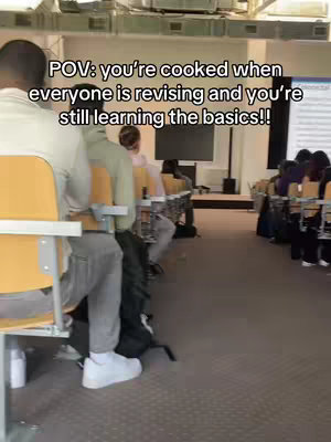 A classroom setting with rows of students facing a screen. The foreground shows the back of a person in a white shirt and grey pants, seated at a desk with a yellow chair. The text "POV: you're cooked when everyone is revising and you're still learning the basics!!" is superimposed on the image.