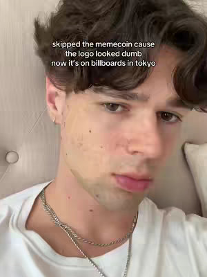 A well-lit, close-up shot of a young man's face. The man, who has slightly tousled, dark brown hair, is looking directly at the camera with a neutral expression. He's wearing a white t-shirt and a silver necklace. The background appears to be a soft off-white surface, possibly a piece of furniture. Text overlaying the image at the top reads, 'skipped the memecoin cause the logo looked dumb now it's on billboards in tokyo'.