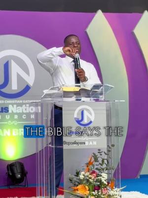 A man in a white shirt and black tie is standing behind a clear podium with a microphone in his hand. The background is a stage with a purple backdrop featuring circular and arched design elements. The hook text is overlaid in a blue box. Decorations in front of the podium include cut flowers.