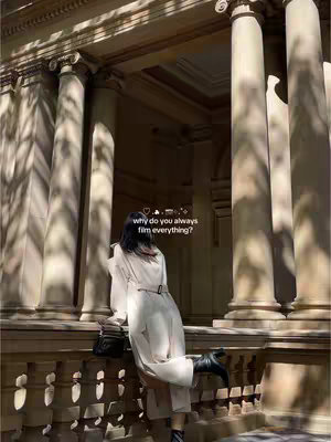 The image shows a person from behind, standing on what appears to be a balcony or ledge with decorative stone architecture in the background. The person is wearing a long, light-colored coat and black boots. The background is a mix of beige stone structures, pillars, with shadows and sunlight, suggesting it's an outdoor location during the day and text overlay that says "why do you always film everything?"