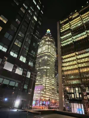 A low-angle shot of a nighttime city scene dominated by tall buildings with lit windows. The central building is a skyscraper with a distinctive illuminated top. The sky is dark, emphasizing the bright lights of the buildings. The composition suggests an urban environment, possibly a financial district.