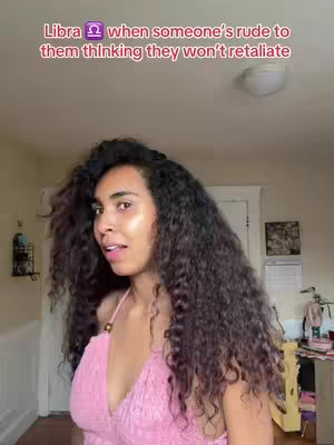 A woman with dark curly hair in a pink top standing in a room, looking slightly to the side. Text at the top reads: Libra ♎ when someone's rude to them thinking they won't retaliate.