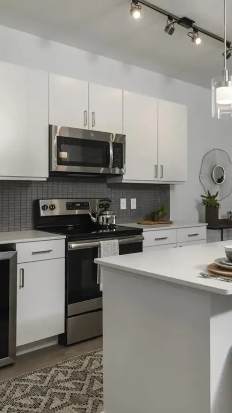 A kitchen with white cabinets and a stainless steel microwave above the stove. A white countertop extends to the right with a round decorative piece on the wall and overhead lighting. The image focuses on the modern kitchen's design.