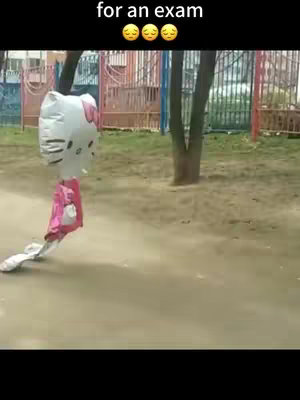 A Hello Kitty balloon with its pink dress is lying on a light brown path. The balloon is facing left, appearing deflated or dragging. The background shows green grass, trees, and a glimpse of buildings with a pink fence.