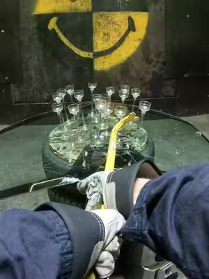The image captures a first-person perspective of someone about to smash a table laden with glasses with a yellow crowbar. The background features a black and yellow smiley face and a damaged wall, while the table sits above a tire. The person's gloved hands and a denim jacket are in the foreground, with the focus on the action.