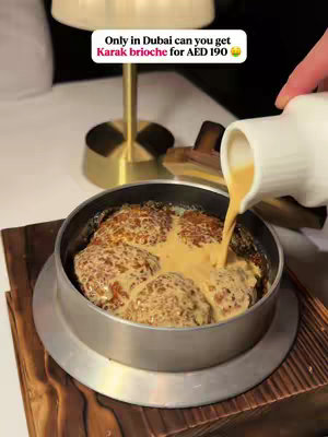 The image shows a close-up shot of a metal bowl containing a baked dish, likely bread, with a yellowish sauce being poured over it from a white pitcher held by an unseen hand in the background. The dish is set on a wooden surface. In the background, there's a mid-century modern style lamp. A text overlay in a speech bubble reads "Only in Dubai can you get Karak brioche for AED 190".
