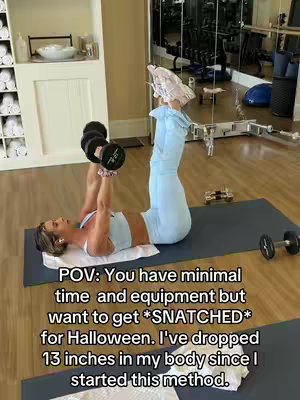 A woman is lying on her back on a blue mat, with her legs raised and dumbbells in her hands, working out her abdominal muscles. The background includes a gym setting with fitness equipment and a mirror.