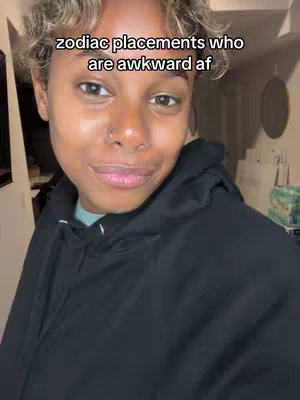A person is centered in a medium shot with a neutral expression, wearing a black hoodie in a casually lit indoor setting. The background is slightly blurred but suggests a residential space. Overlayed text states "zodiac placements who are awkward af".