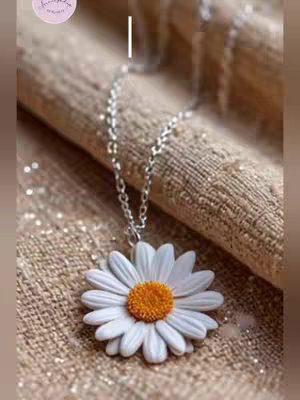 The cover image features a close-up of a daisy pendant. The pendant is centered; the daisy has white petals with an orange center, supported by a silver chain. It rests on a textured, beige surface in the background, possibly fabric or a rug. A logo or watermark is visible in the top left corner. The image is well-lit, highlighting the pendant's details and the texture of the background.