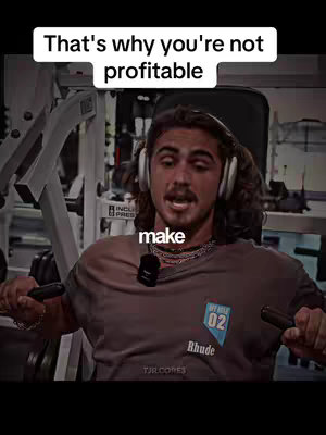 A man is working out at the gym. The video cover image shows a man wearing a gray shirt with the phrase "That's why you're not profitable" written in a white rectangle at the top. The man is wearing the headphones and a microphone, the background has a blurred gym setting with gym equipment visible.