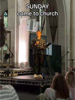 A man in a plaid shirt stands on a stage in a church, holding a microphone and reading from a book. The image is a wide shot, showing the church interior, with text overlaying the top.
