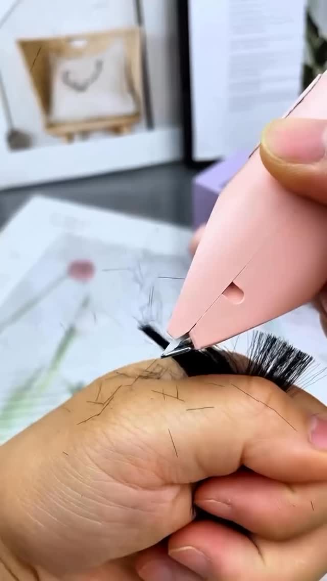 A close-up shot shows a hand holding a pink electric grooming tool, cutting hair against a dark hand. The tool is positioned just above the base of an eyelash, with trimmed hairs floating. A white background with blurred decorative objects is visible in the background.
