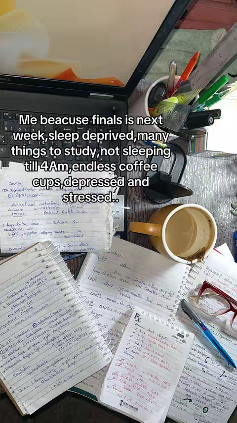 A cluttered desk filled with open notebooks, a laptop, pens, and a mostly-empty mug of coffee. Handwritten notes and study materials are scattered across the surface, suggesting a study environment. The laptop is open with a visible keyboard and screen reflecting the overhead light.