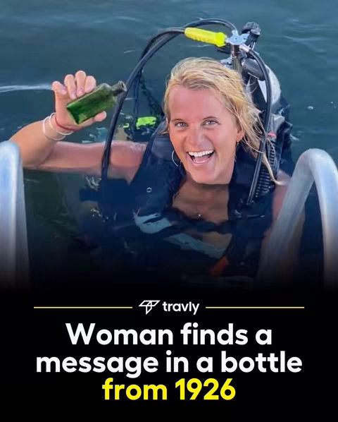 The image features a woman in a diving suit submerged in water, holding a small green bottle. She is smiling widely, with a background of water and the diving equipment she is using. The lower portion of the image features a text overlay in white and yellow font, stating 'Woman finds a message in a bottle from 1926'.