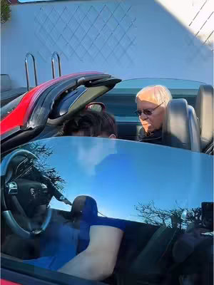 A photo shows two people inside a red convertible, with the top down. The primary focus is on the person in the passenger seat, an older individual wearing sunglasses. The other person is partially visible in the driver's seat with their face obscured. The car is parked outside on a sunny day, reflected in the windshield, a blue sky can be seen behind.