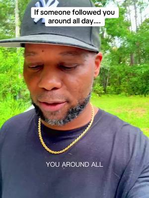 A man with a black baseball cap and a black shirt with a gold chain is looking at the camera with trees in the background.