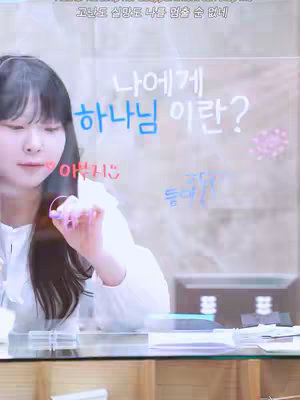 A woman is seen in front of a transparent surface, upon which text and drawings have been added. She is seated at a table, her features soft, and she is holding a pen near the surface as if about to write. The background is blurred, suggesting a room or office setting.