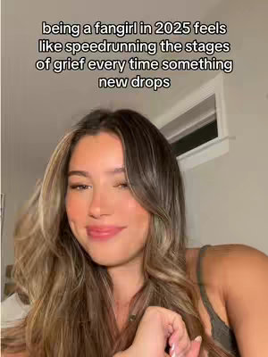 A young woman with long, wavy brown hair is shown in a selfie-style shot. The background is a softly lit room, with a window in the back right and a hint of a wall on the left. The woman is facing the camera, her expression gentle and inviting, with a slight smile. She wears a casual top and the overall composition is warm and natural, conveying a sense of intimacy or casual conversation.