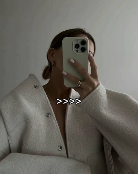 A woman is taking a selfie with a light-colored phone. The background is a plain, neutral wall. She is wearing a light-colored jacket, and the phone is obscuring the majority of her face. Above her jacket, the text ">>>>" is present.