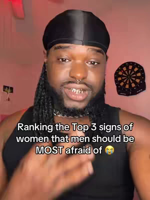 The cover image is a selfie-style video. The creator, a Black man with braids, a black durag, and gold teeth, is centered in the frame with the background featuring a pink-toned wall and a dartboard. Text overlay at the bottom reading: "Ranking the Top 3 signs of women that men should be MOST afraid of 😳"