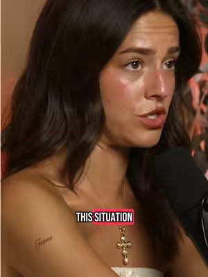 A woman with long dark hair is facing the camera and talking. The image shows a close-up view of her face, focusing on her expression and the area around her mouth, neck and shoulder. She  is wearing a white strapless top and a gold cross necklace. A tattoo is seen on the arm.