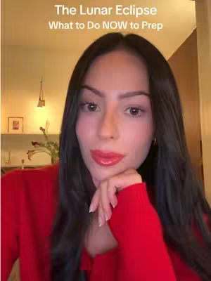 A woman with long dark hair and red lipstick is shown in a close-up shot. She's wearing a red top, and her right hand is touching her chin. The background is softly blurred, with some lighting and decor visible.
