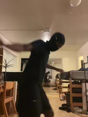 A person wearing all black and a black face mask is standing in a well-lit room with an open floor plan. The person is in motion, possibly dancing or moving enthusiastically, with arms outstretched. The background shows furniture and other typical household items. The overall composition suggests a casual indoor setting.