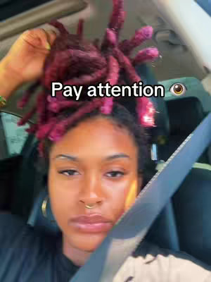 A selfie shot from inside a car, featuring a Black woman with maroon-colored locs styled upwards. She has a gold nose ring and hoop earrings. The background suggests a sunny day, with visible car details and the driver's seat visible.