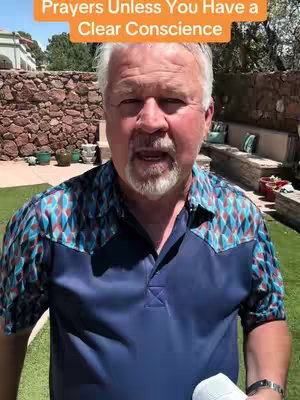 A middle-aged man with white hair and a beard is speaking directly to the camera. He's in a well-lit outdoor setting, wearing a patterned shirt, and the background includes a stone wall and greenery.
