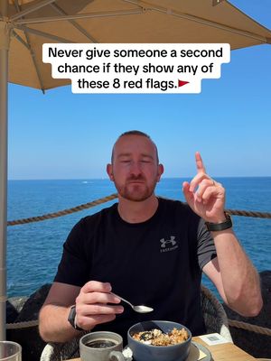 A man with a beard sits at a table with food, pointing upwards with his right index finger. The background shows a bright blue sky and a view of the ocean.