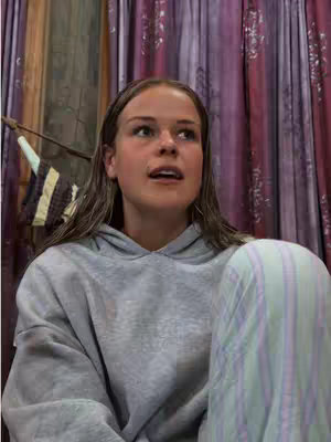 The cover image features a young woman with light brown hair, wearing a gray hoodie and striped pajamas. She is positioned in front of a backdrop of purple and pink curtains. The overall composition is a close-up shot, focusing on the woman's upper body and facial expression.