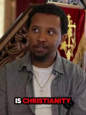 A man with dark hair and stubble is centered in the frame. He wears a gray and white checkered shirt over a white t-shirt and a cross necklace. Behind him, a red draped backdrop with gold religious symbols is partially visible. The lower portion of the screen holds a black bar with white text.