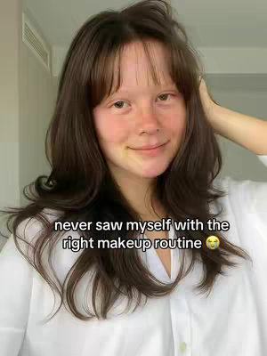 A selfie showing a young woman with long, wavy brown hair and bangs. She is wearing a white shirt and has a soft smile. Text overlayed at the bottom says, "never saw myself with the right makeup routine." The background is slightly blurred, suggesting an indoor setting.