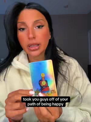 The cover image features a woman holding a tarot card. The woman is fair-skinned with long dark hair pulled back. Her makeup is visible, with defined eyebrows, and shaded lips. The tarot card is held in front of her chest, the image has a bluish background with an image on it. Below the image is a black rectangle that contains white text.