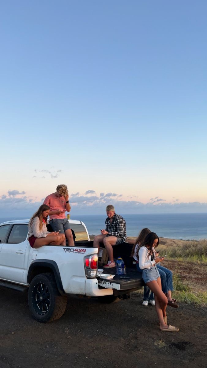 The cover image shows a group of five young adults posing on the tailgate of a white pickup truck parked on a dirt road, overlooking a coastal scene at dusk. The sky is a gradient of blue and pink hues, suggesting sunset or sunrise. The individuals, a mix of male and female, are casually dressed and are using their phones. The truck bed is filled with gear, hinting at a road trip or outdoor adventure. The composition is balanced, and the color palette creates a warm, inviting atmosphere.