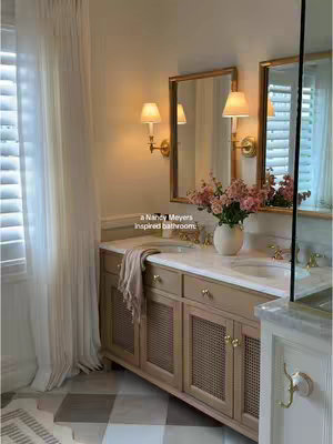 A well-lit bathroom with a vanity, mirrors, and elegant decor. The vanity is light brown with woven cabinet fronts and brass hardware. Above is a marble countertop of two sinks, which is framed by two wall-mounted mirrors framed in gold. Soft, sheer curtains are seen to the left of the sink in a window. There are also flower arrangements and decorative lighting, adding to the overall aesthetic.
