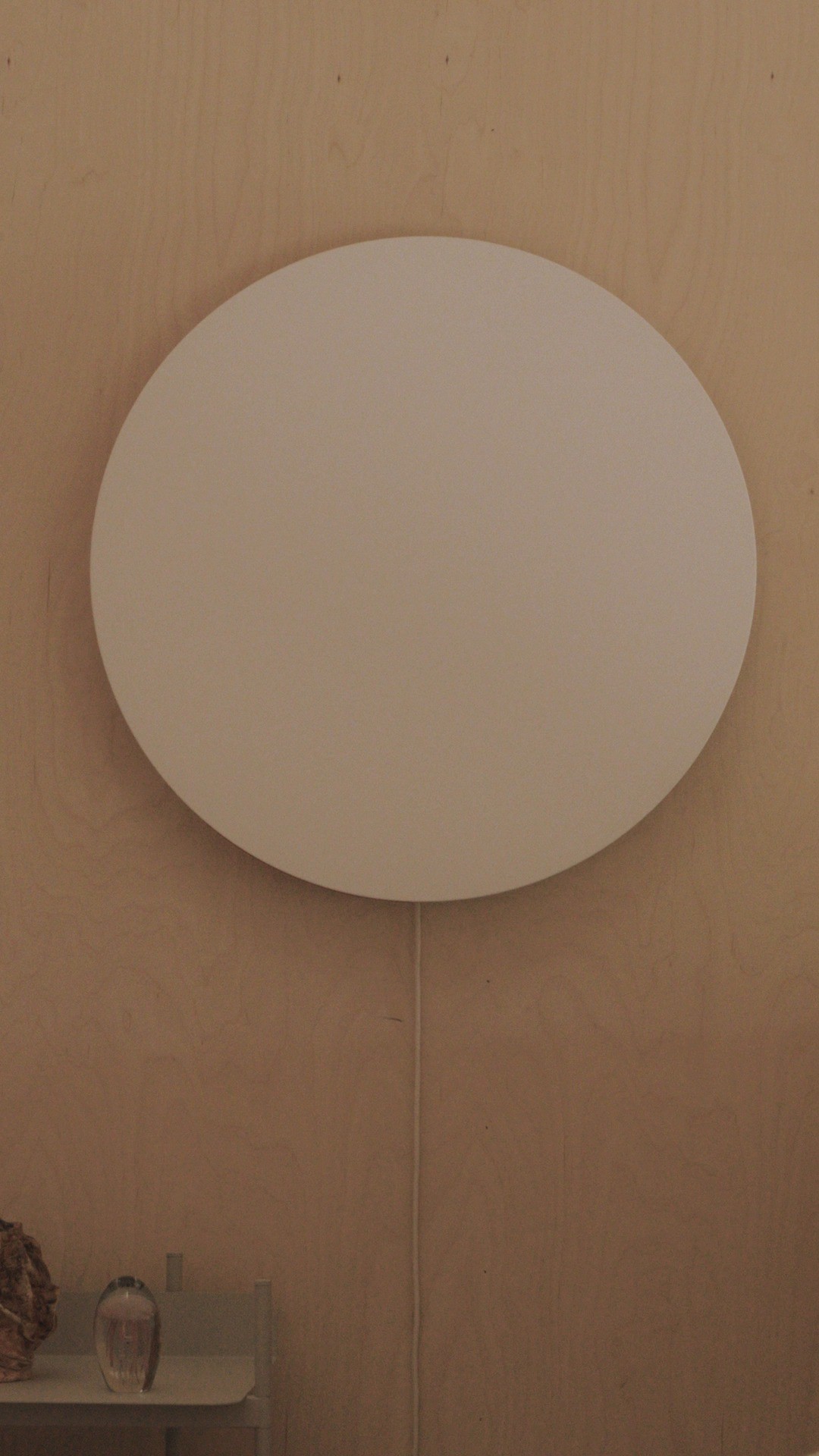 The image shows a matte white, circular wall lamp mounted on a light brown, wooden panel. The lamp has a simple design with a circular shape and a thin electrical cable hanging down. The background is a solid, slightly textured wall that complements the lamp's clean aesthetic, while a few objects are visible in the lower left corner.