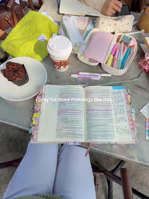 An overhead shot of a table laden with items suggestive of a morning devotion. An open Bible is in the center, with notes and tabs. A cup of coffee, a plate with a muffin, pens, and other stationery items are scattered around. The composition is bright and airy, with a soft focus.