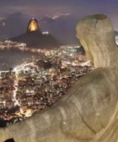The image shows a night view overlooking the city from the vantage point of the Christ the Redeemer statue in Rio de Janeiro. In the foreground, the statue is visible in tan, with the city lights and Sugarloaf Mountain in the background. The overall composition provides a serene, inspiring view. The lighting is somewhat dim, but gives a warm effect reflecting a spiritual theme.