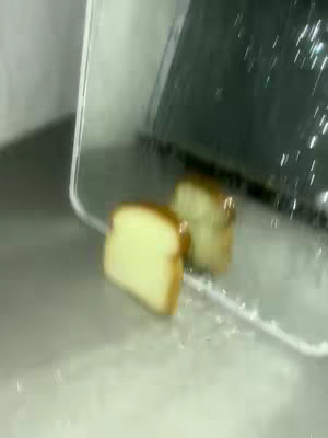 A slice of bread is falling into a clear container, possibly a toaster. The bread has a light brown crust and a pale yellow interior. The container reflects the bread piece. The background is unclear.