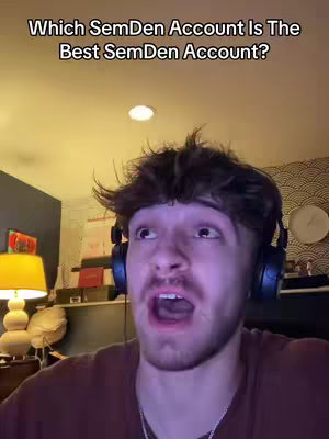 A selfie-style shot features a young man with short, spiky brown hair and a slight beard, his expression one of surprise or slight shock with his mouth open. He wears a dark headset around his neck and is in a room with a warm, orange-yellow lighting. The background has several soft elements, including a lamp, a decorative piece, and a clock. The text 'Which SemDen Account Is The Best SemDen Account?' is displayed above the man’s head to indicate the topic.