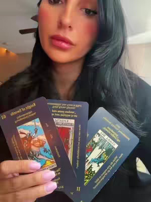A woman with long black hair is holding three tarot cards, she is looking at the camera. She is wearing a black top and has pink blush on her cheeks and pink lipstick. The background is a light-colored surface.
